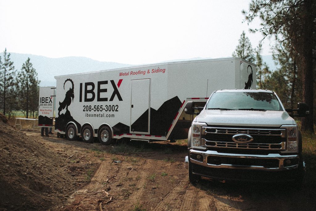 Large white IBEX trailer with 'Metal Roofing & Siding' text at a construction site, towing a Ford truck in the foreground, dirt ground and trees in background