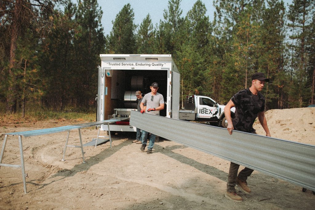 Workers unload a long metal beam from a trailer at a sandy outdoor construction site with trees in the background.